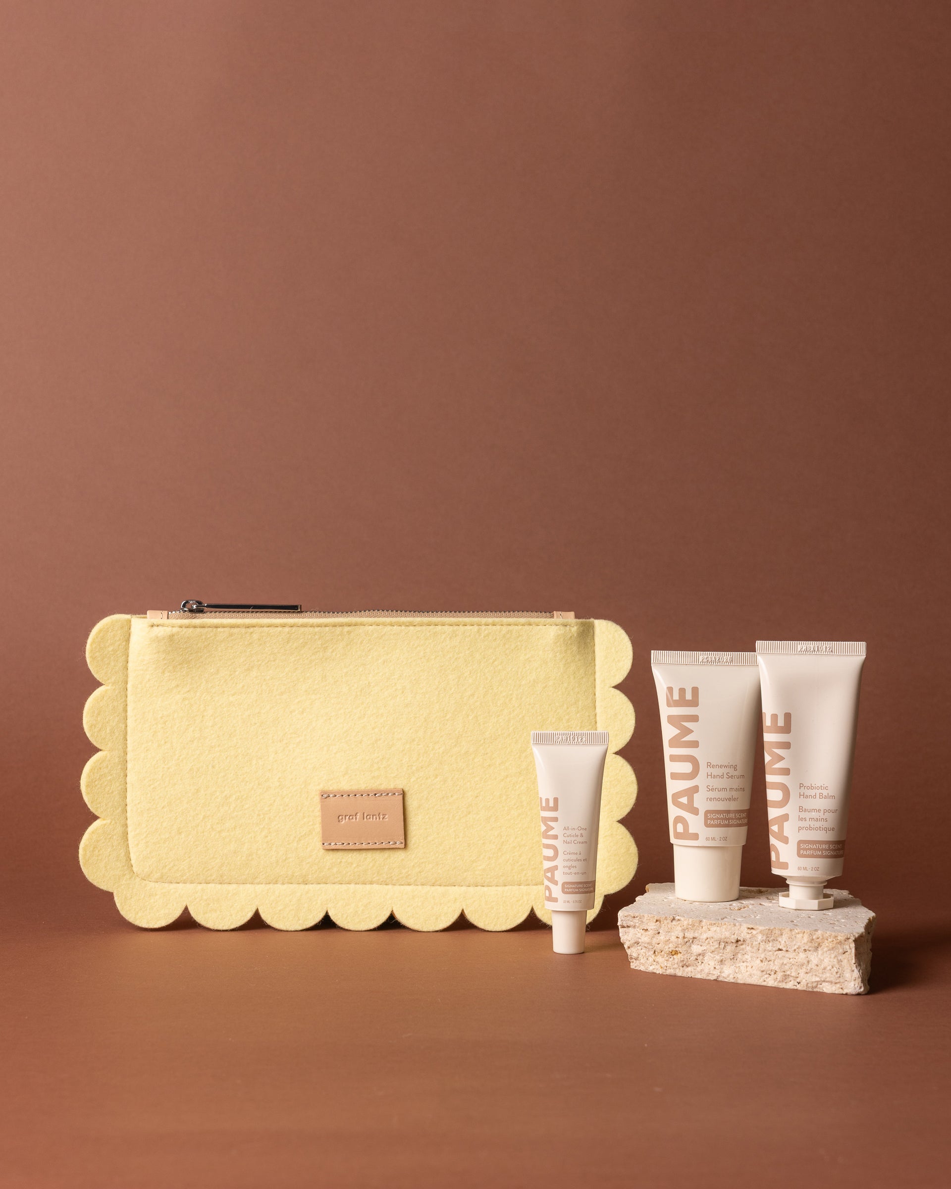 A yellow Graf Lantz Merino Wool Pouch sits beside four PAUME Hand Care Set tubes of different sizes, all arranged on and around textured beige stones with a brown background.
