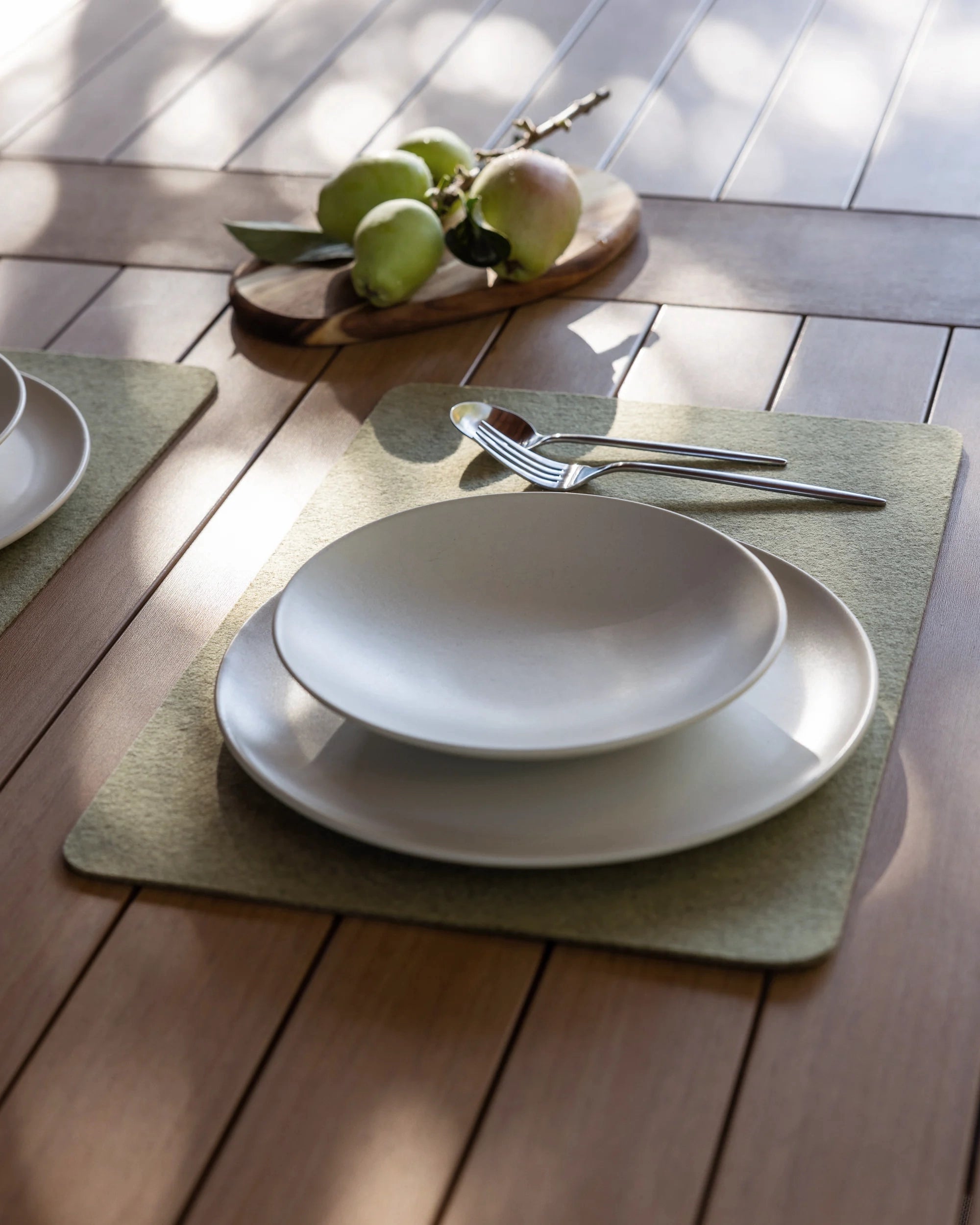 A wooden table set features a white plate, bowl, fork, and spoon on a heat-resistant graf lantz Merino Wool placemat. In the background, a small wooden board with apples and a branch casts soft shadows over the elegant setup.