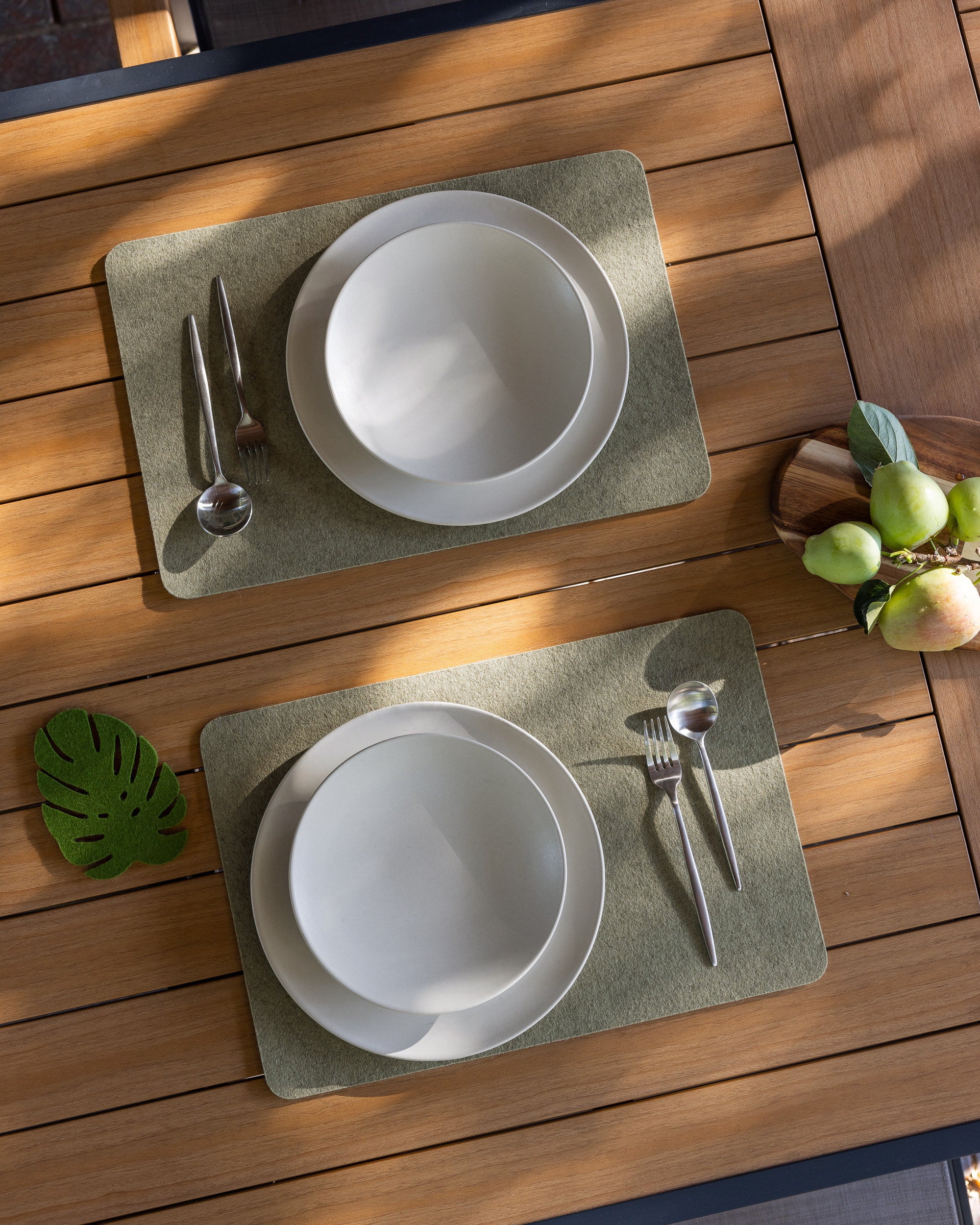 Two place settings on a wooden table feature graf lantz merino wool placemats, each holding a white bowl and plate, fork, knife, and spoon. A small cutting board with green and ripe pears is on the right. A leaf-shaped coaster rests nearby, adding charm to the cozy dining setup.