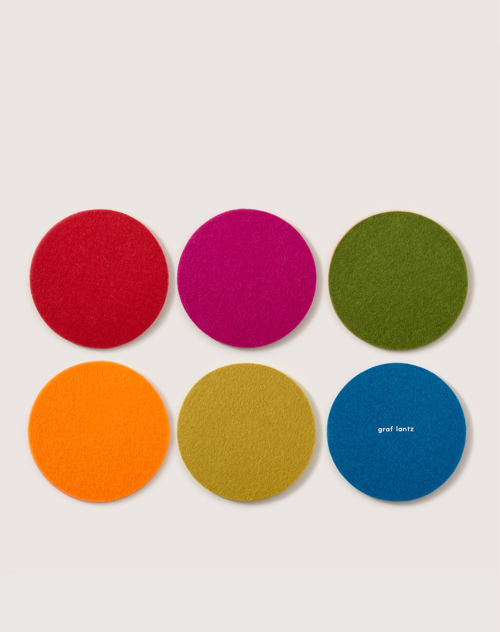 Six round coasters made from soft German Merino wool are arranged in two rows: red, pink, green on top; orange, yellow, blue on the bottom. The blue coaster features graf lantz against a light gray background.