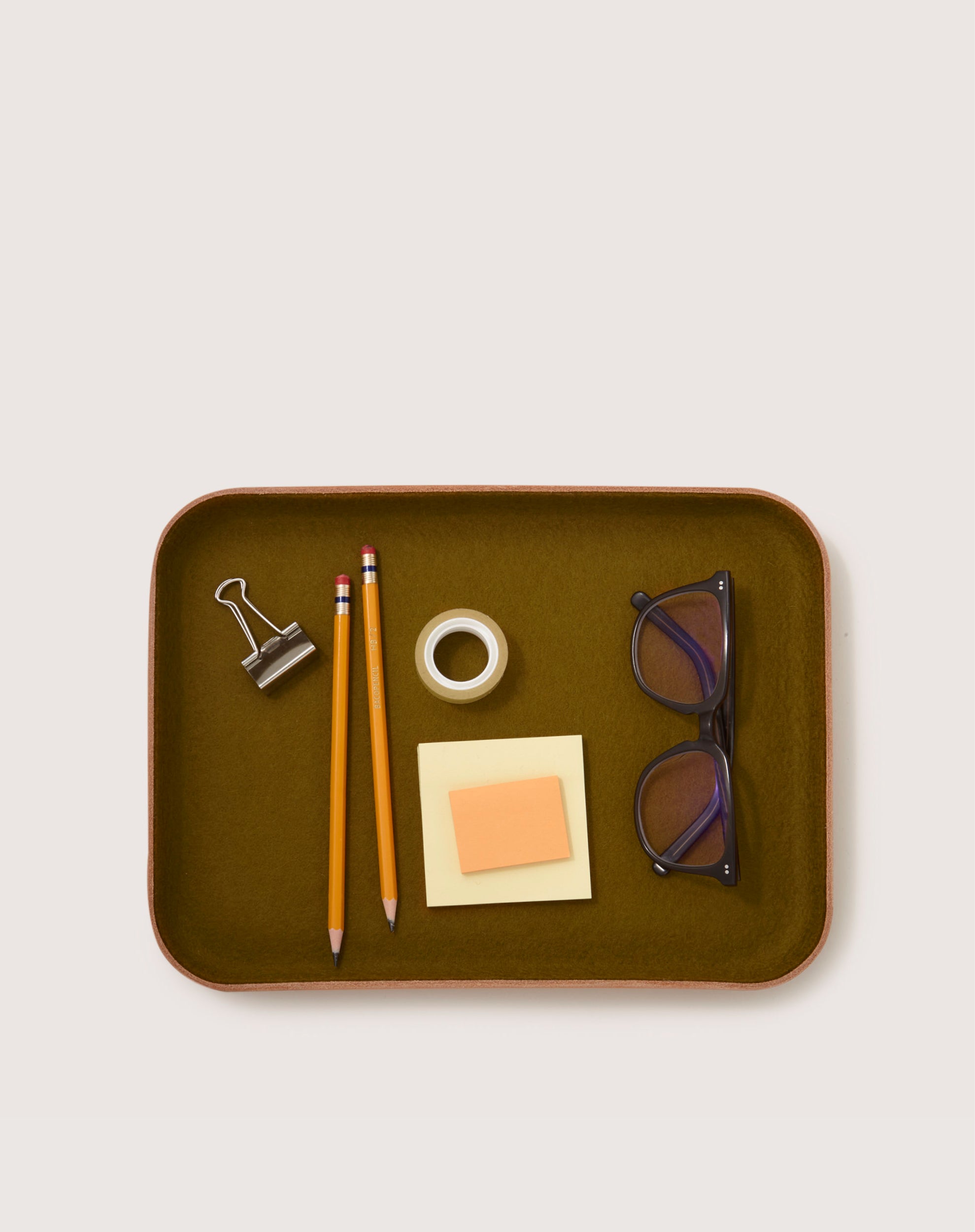 The graf lantz catchall tray holds two yellow pencils, a binder clip, a roll of tape, sticky notes, and glasses. It features a rectangular design with a muted olive-green interior that complements its sculpted leather finish.