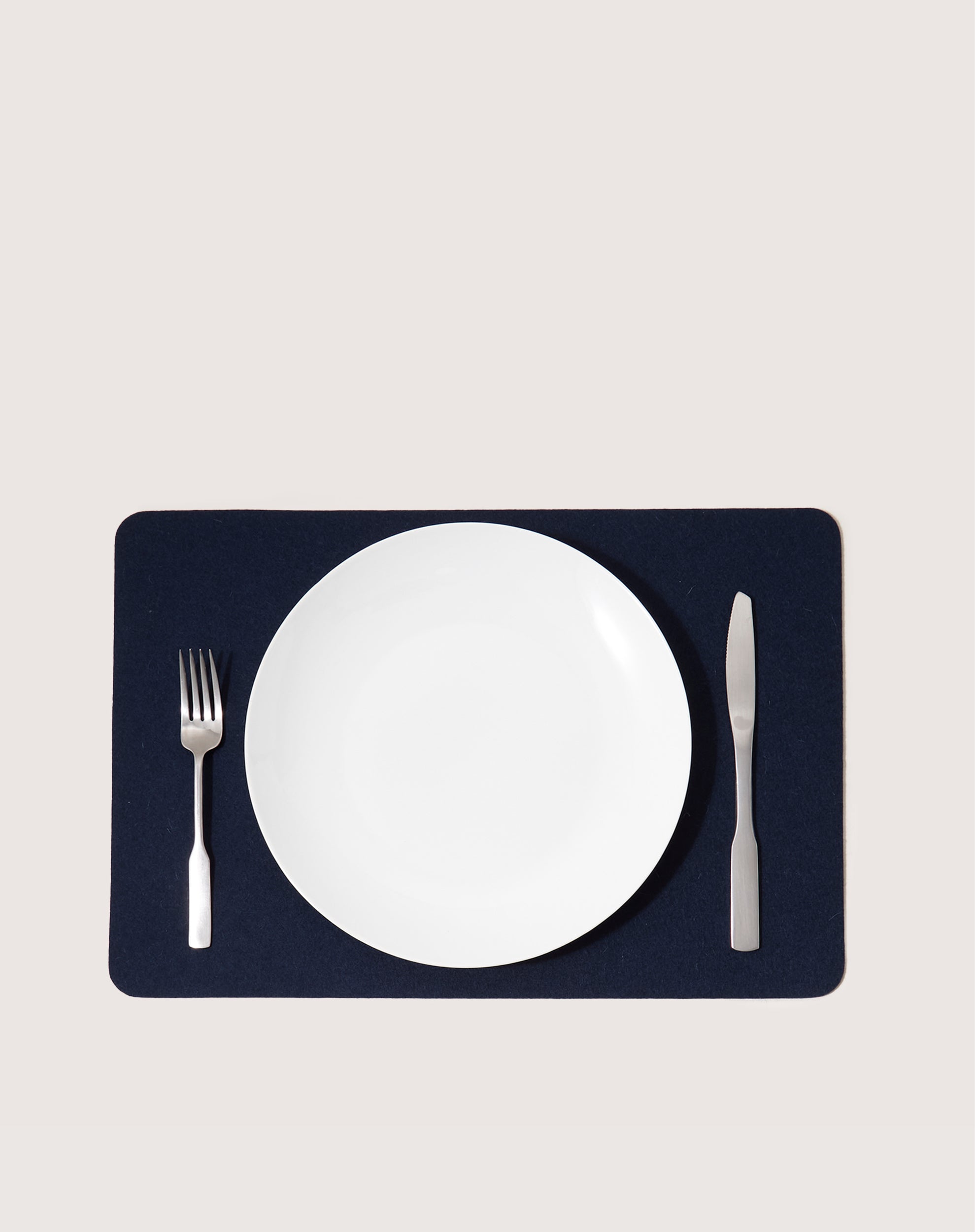 A white plate is centered on a dark navy graf lantz Merino Wool Placemat, valued for its heat-resistant and moisture-wicking features. A fork lies left of the plate, with a knife to the right. The background is light gray.