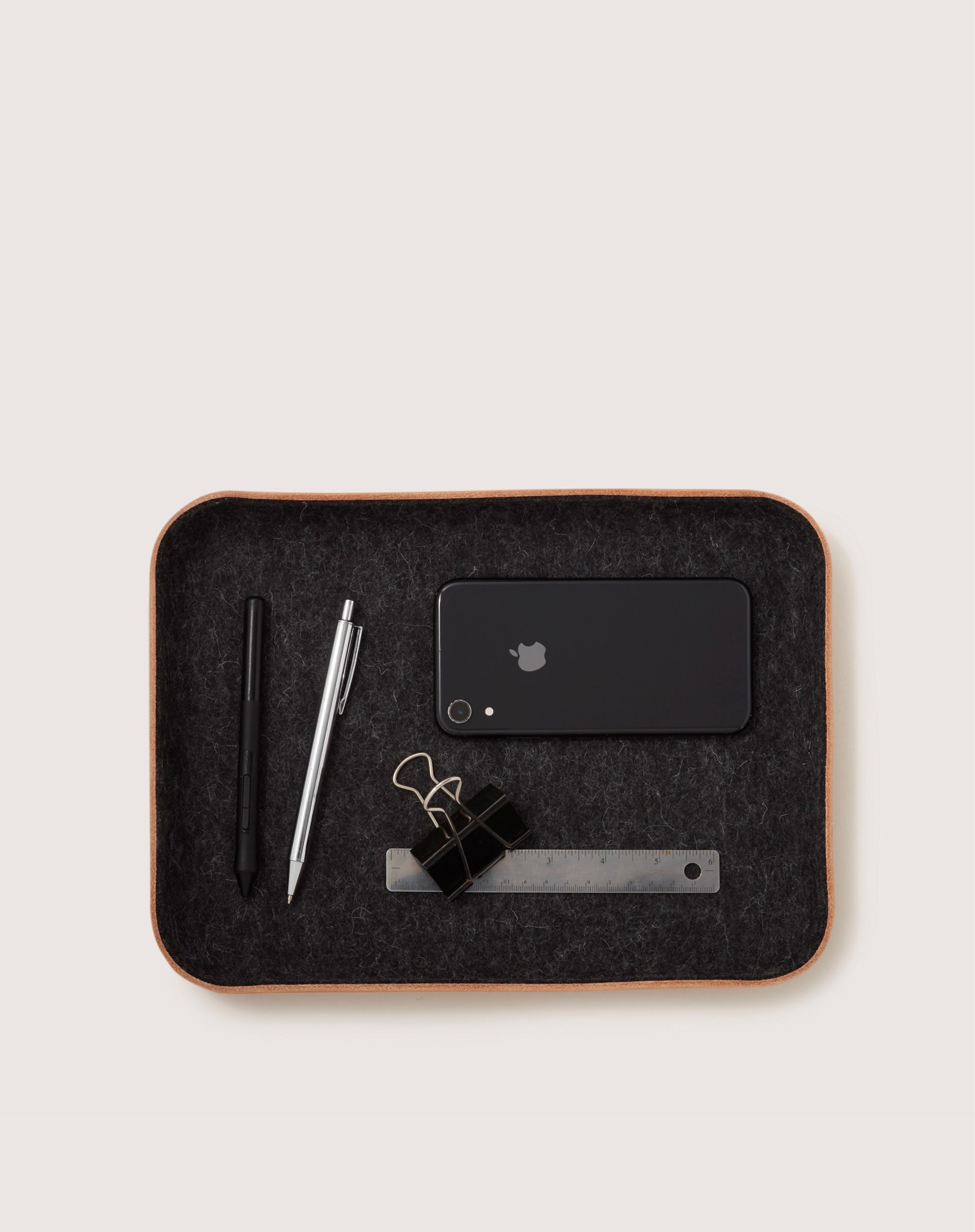 A graf lantz Merino wool catchall tray holds a black smartphone, two pens, two black binder clips, and a metal ruler against a neutral background.
