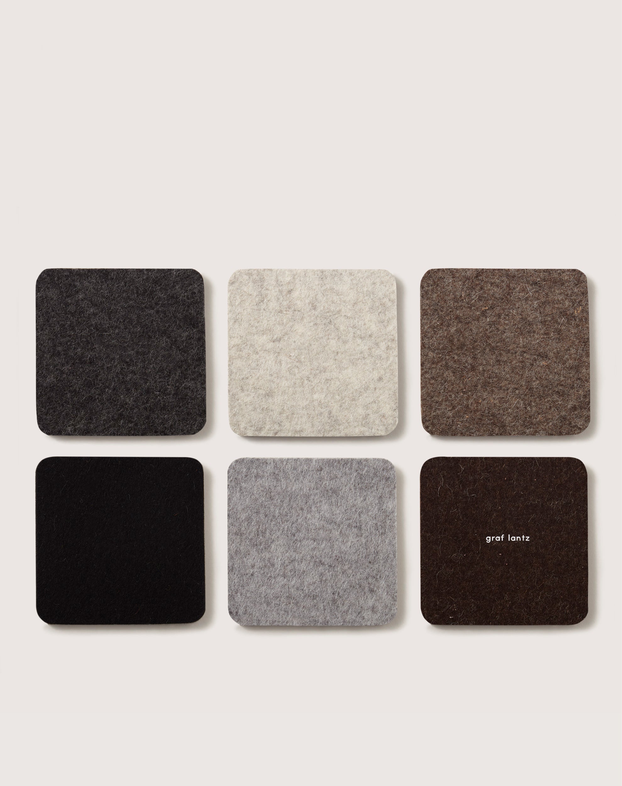Six square felt coasters from graf lantz, crafted from Merino wool, are arranged in two rows. The top row displays dark gray, cream, and brown; the bottom features black, light gray, and dark brown with graf lantz branding.