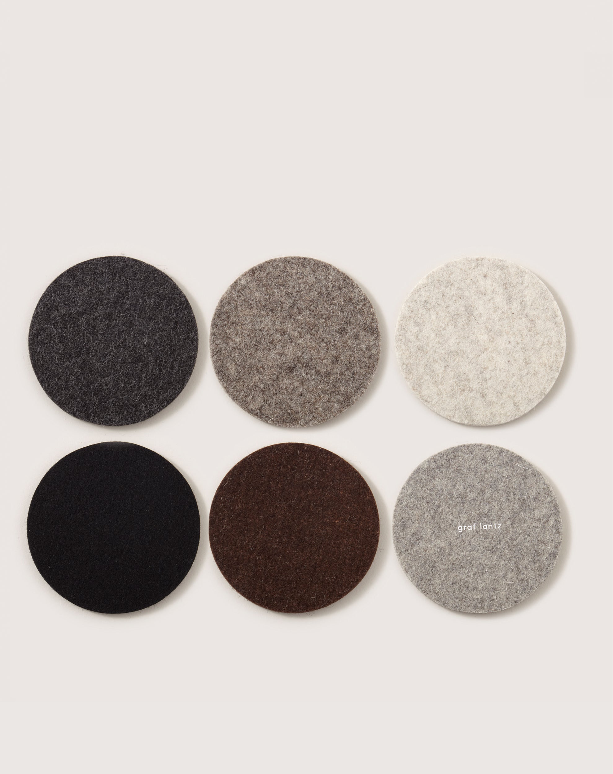 Six round coasters made from German Merino wool in shades of gray, brown, and black are arranged in a grid on a light background. The middle right coaster features the text graf lantz, adding an elegant touch to your home décor.