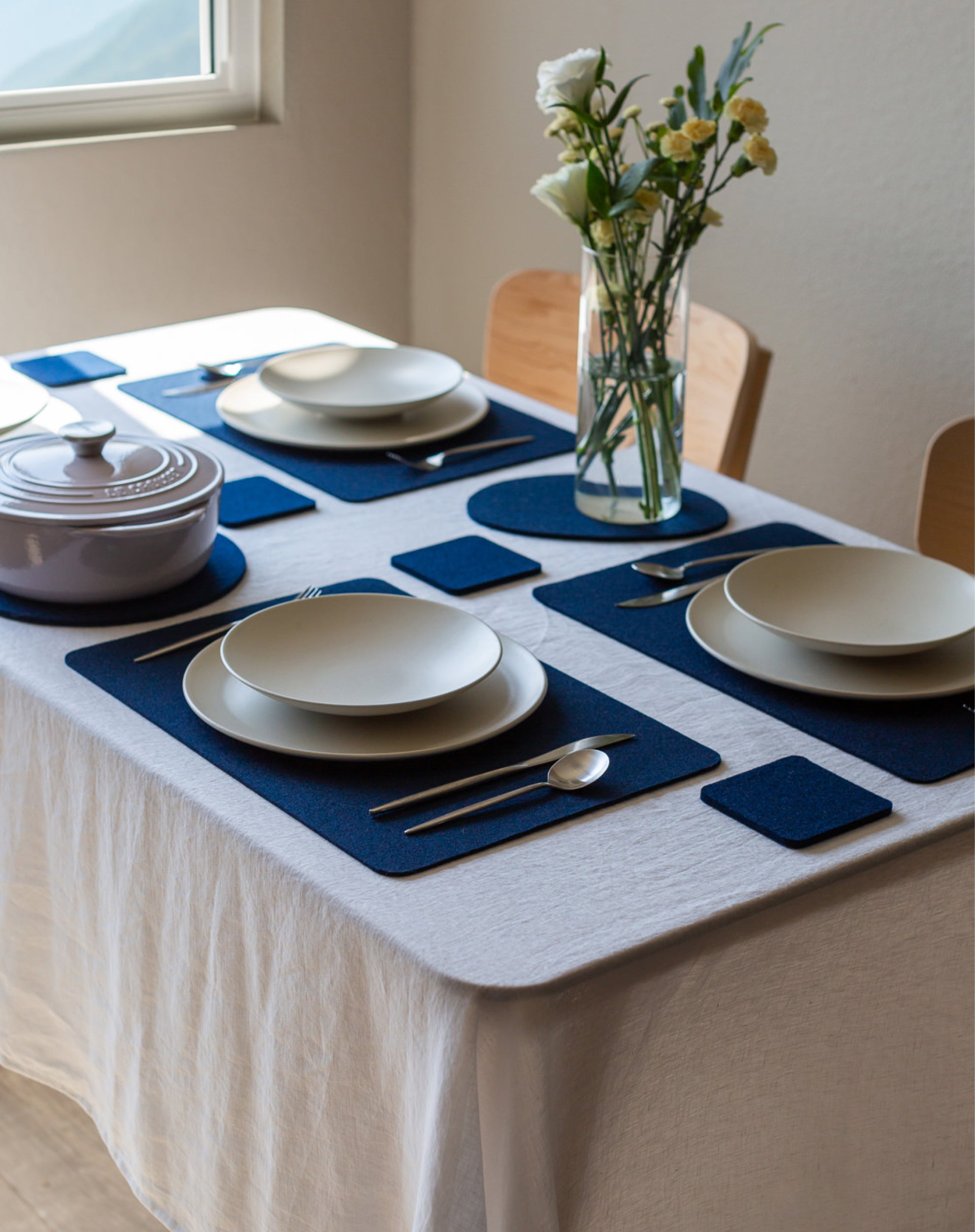 A neatly set dining table with a white cloth features four place settings, each with a graf lantz Merino Wool Placemat, white plates, silver cutlery, and covered dish. A vase of white and yellow flowers by the window adds charm to the elegant setting.