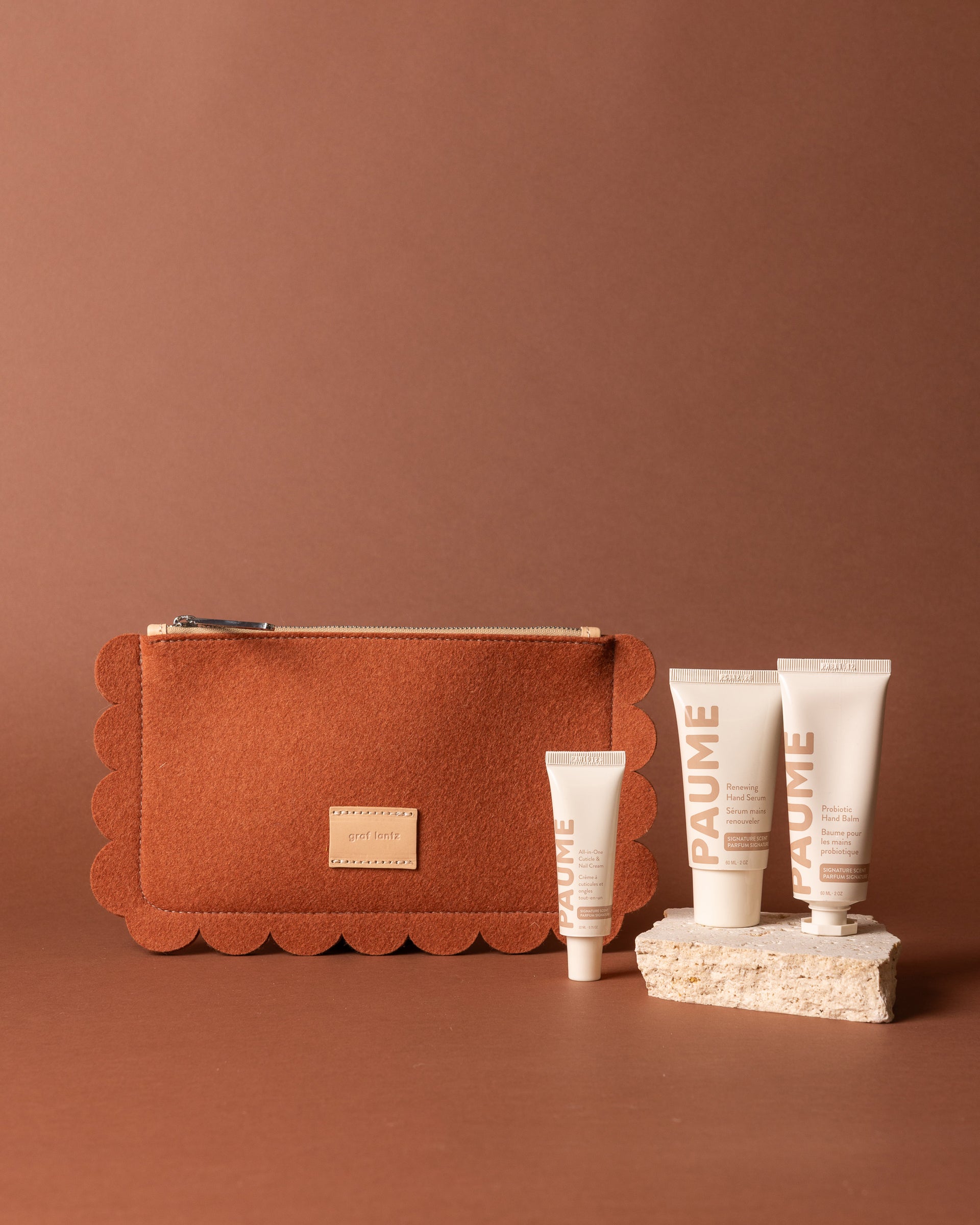 A brown Graf Lantz Merino Wool Pouch is placed beside four white PAUME skincare tubes, including Paume Hand Serum and Probiotic Hand Balm, three on a textured stone and one on the surface, against a brown background.