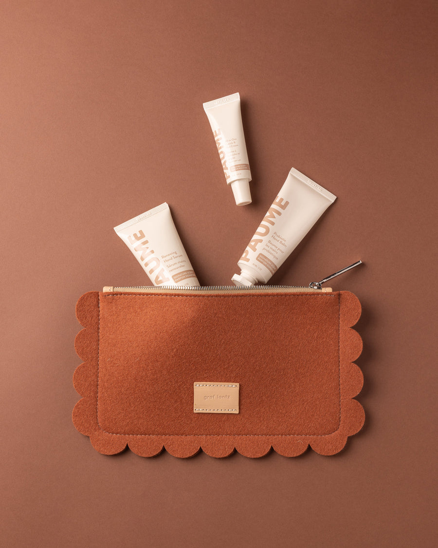 A scalloped-edge brown Merino Wool Pouch by Graf Lantz with a zipper is open, revealing three beige skincare tubes labeled PAUME partially emerging against a matching brown background.