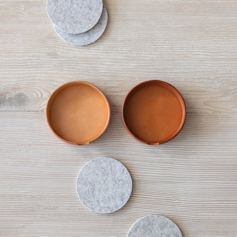 Two vachetta leather coaster holders, the left coaster holder is new while the other showcases patina. Gray Merino wool coasters sitting decorative next to the coaster holders on wooden tabletop.