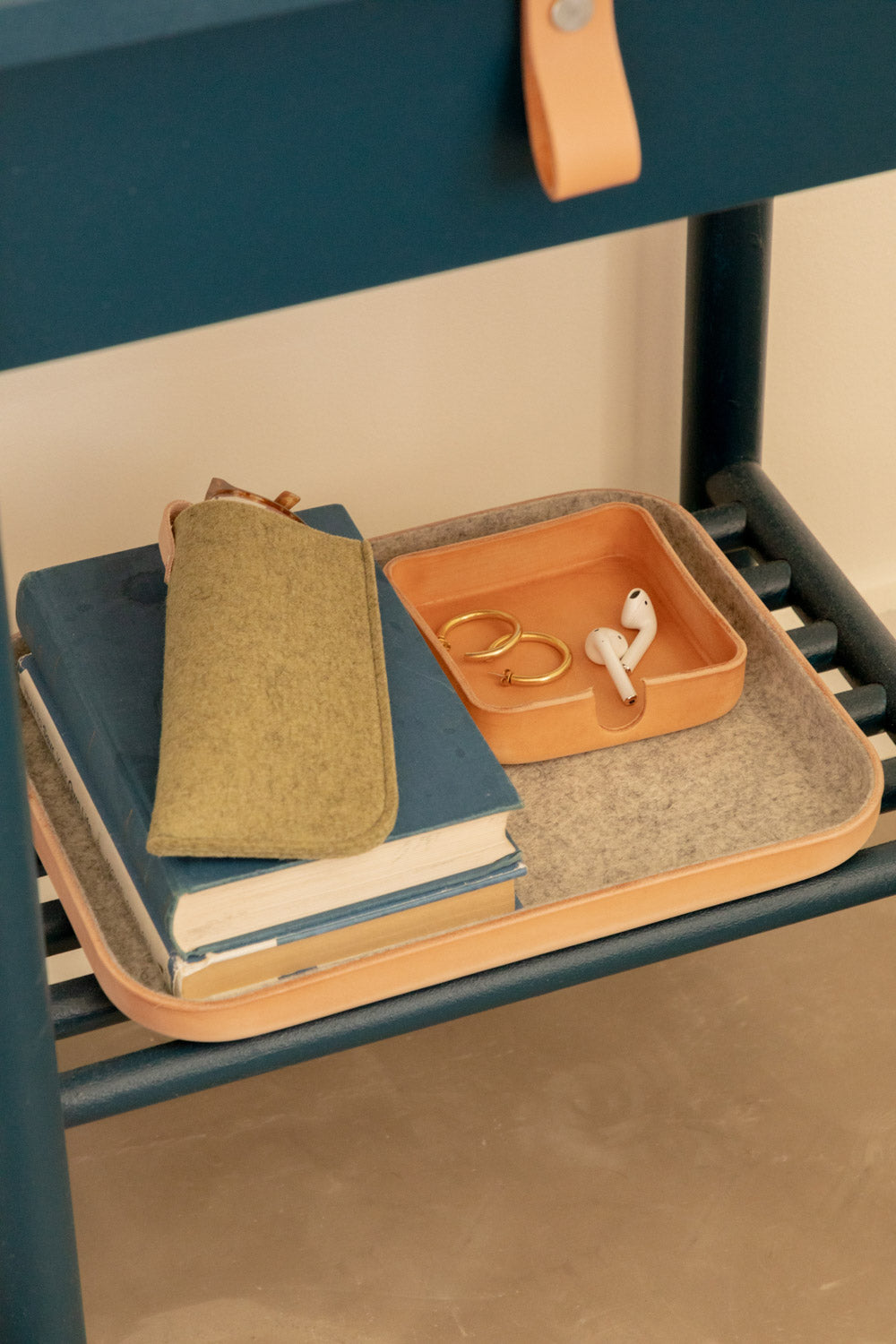 A Kawabon Tray on a dark teal shelf holds hoop earrings, wireless earphones, and a green fabric pouch. Stacked blue books enhance the look against a light-colored wall. Tan accents complete this stylish arrangement, branded by graf lantz.