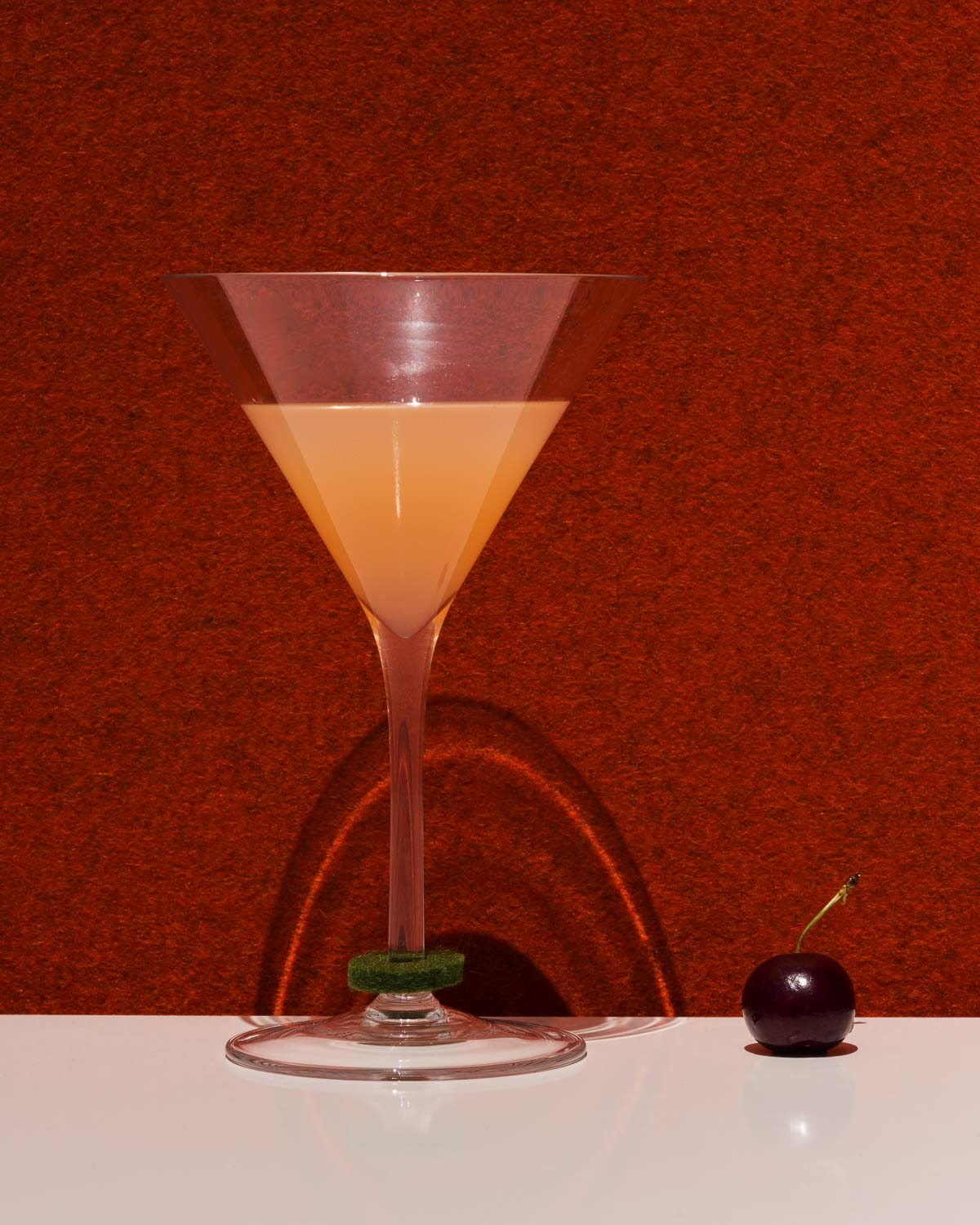 A tall, triangular graf lantz cocktail glass containing a peach-hued drink sits on a white surface against a bold red backdrop. wine glass markers and a single cherry nearby create clear shadows.