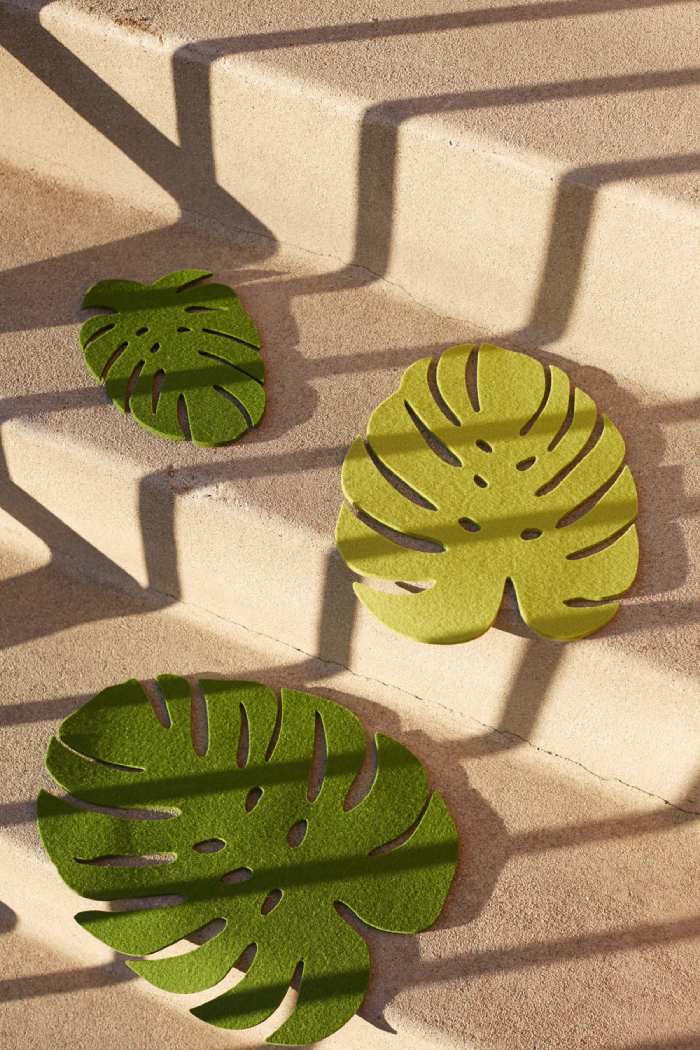 Three eco-friendly, felt monstera leaf cutouts from graf lantz in green adorn beige steps. Shadows from the railings form striped patterns across the steps and leaves, which vary in size to add depth and visual interest.