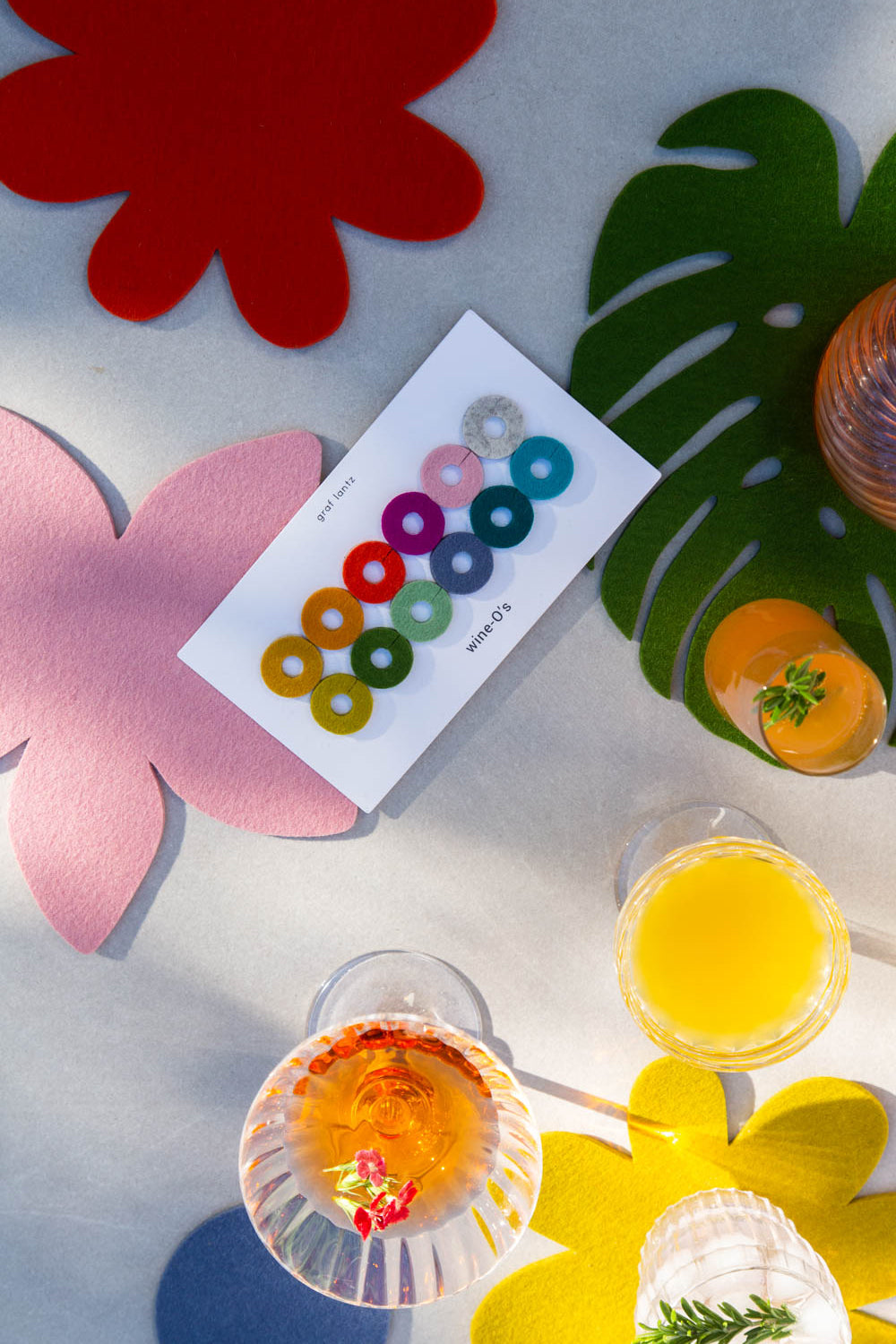 A table features graf lantz’s eco-friendly, felt leaf-shaped placemats and drinks alongside a white card with colorful rings, possibly for Wine-Os markers. The sustainable wool design casts charming shadows, enhancing the elegance.