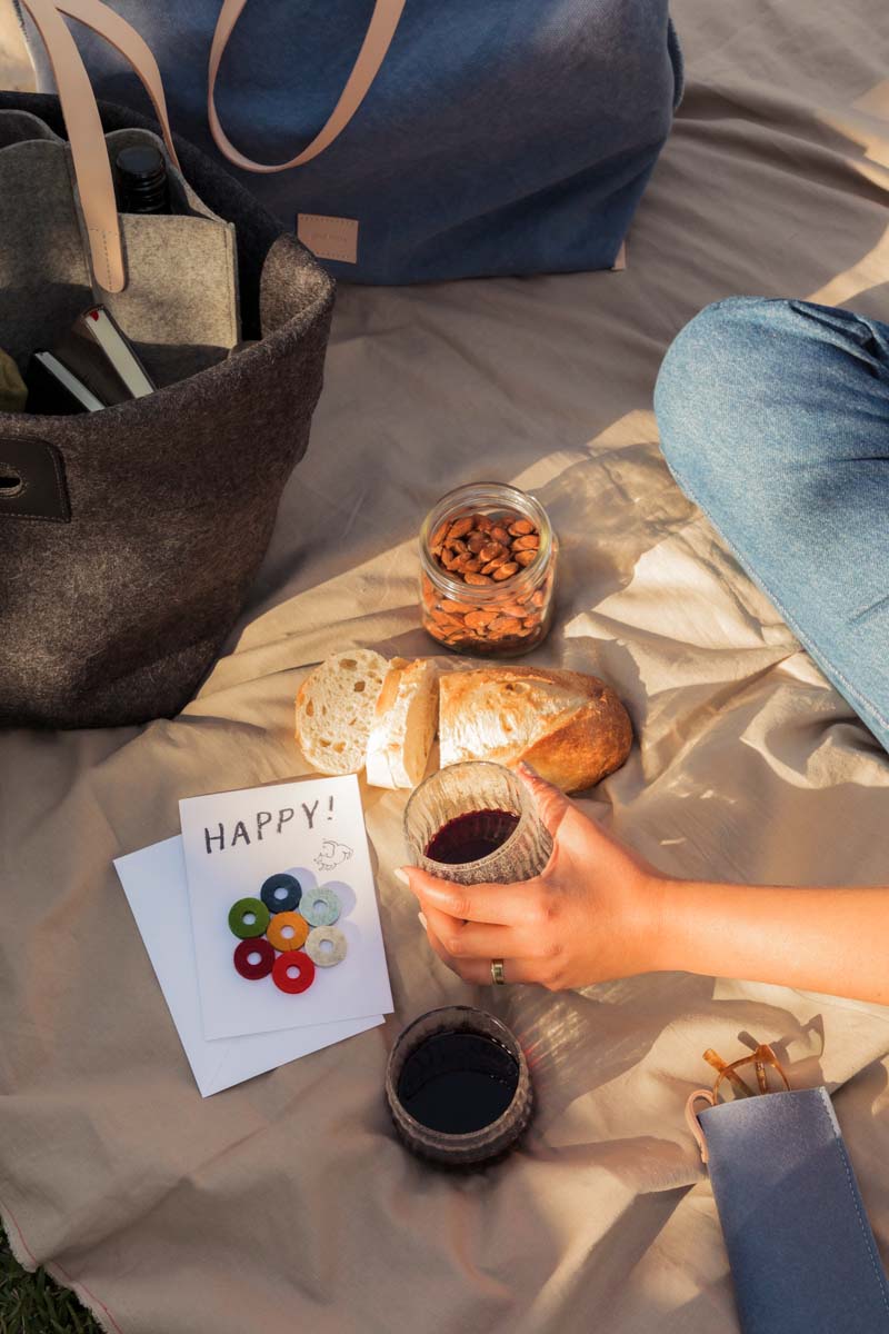 A picnic unfolds with someone holding a glass of red wine. Sliced bread, almonds in a jar, and another filled glass rest on the blanket. Beside them, a tote bag and graf lantz Wine Marker Note Cards displaying HAPPY! in vibrant hues add charm to this delightful scene.