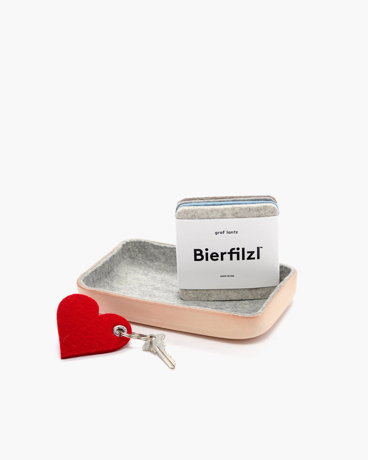 A beige Kawabon catchall tray by graf lantz holds gray Merino wool Bierfilzl coasters. Next to it lies a key with a red heart-shaped keychain, ideal for a housewarming gift. The backdrop is plain white.